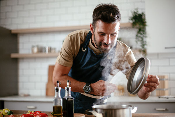 A man in an apron lifts the lid off a pot of steaming food.