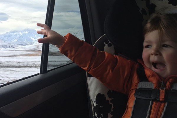 Child in an orange jacket reaches out of car window towards snowy mountains.