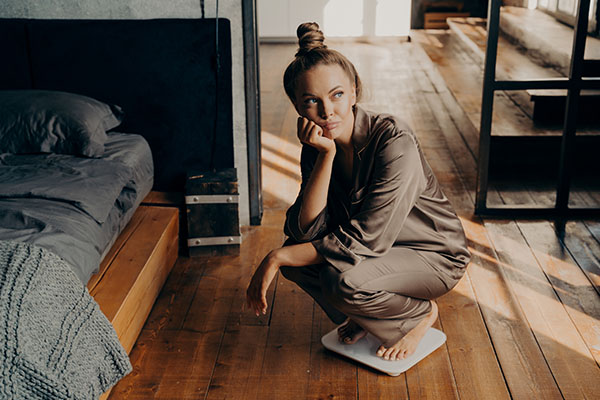 A woman in pajamas sits on a scale in a bedroom, looking thoughtful.